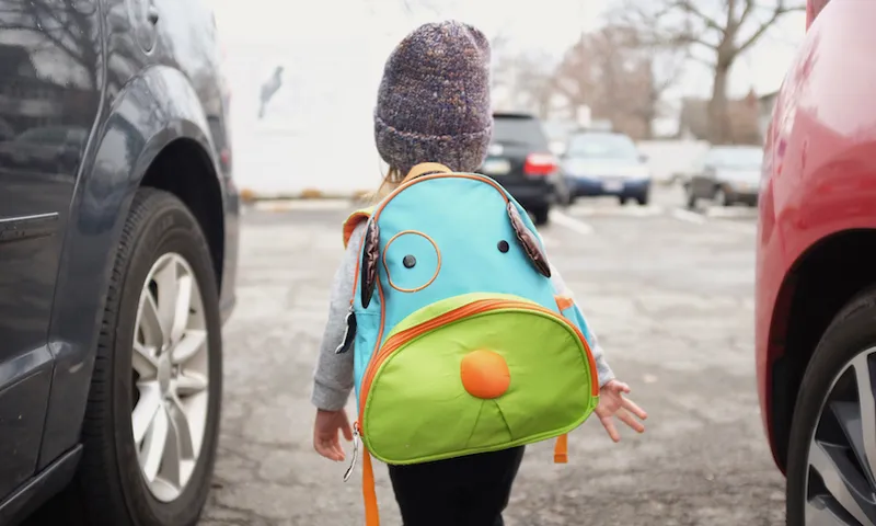 Rear view of young girl carrying bright green and blue backpack, walking in between parked cars.