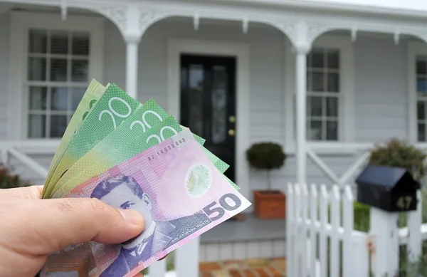 A mans hand holds NZ dollar bills against a front of North American hous.