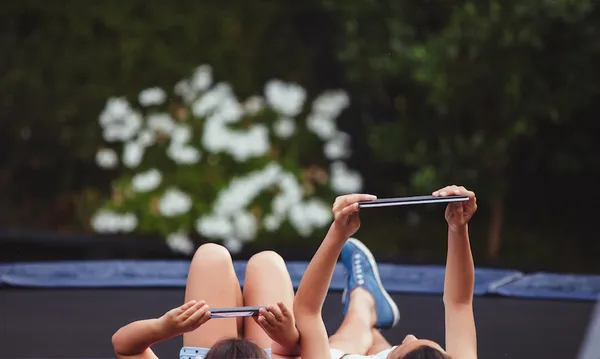 Two young sisters on trampoline playing on tablets.