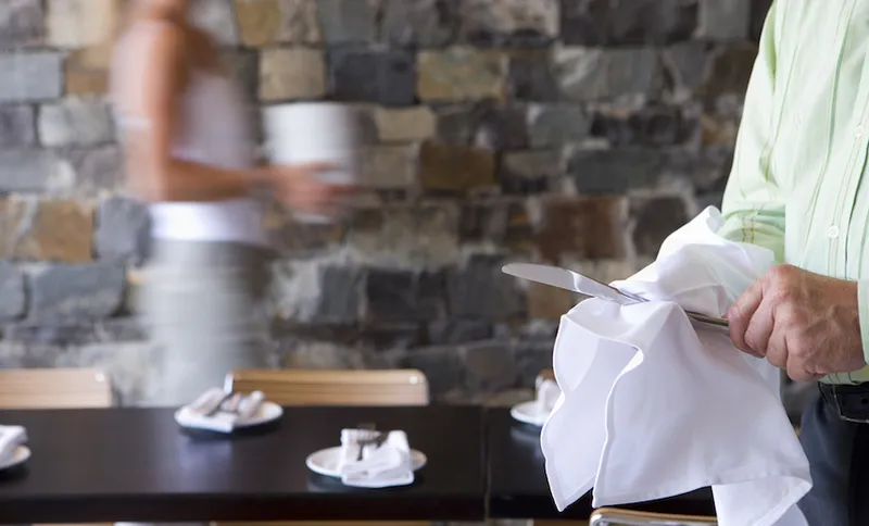 Waiter and waitress laying tables in restaurant, focus on man wiping knife in foreground, side view (blurred motion)