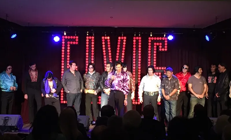 Photo of a line-up of Elvis impersonators on stage, huge neon ELVIS behind them