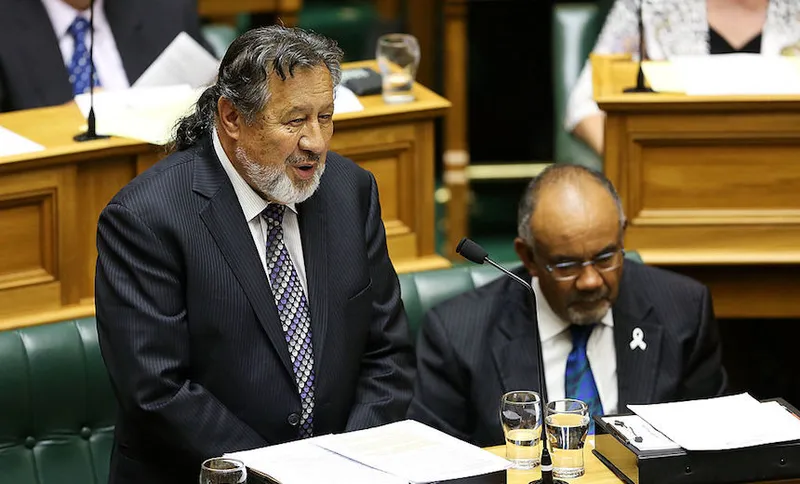 A man in a suit with a grey beard speaks at a podium in a parliamentary chamber, while another man sits beside him, also dressed in a suit, with papers and glasses of water on the desk.