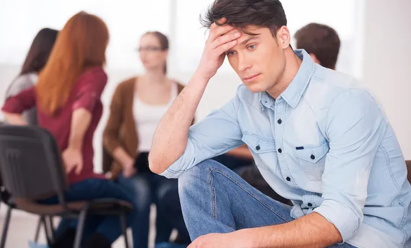 Feeling pain and depression. Depressed young man sitting at the chair and holding head in hand while other people communicating on background