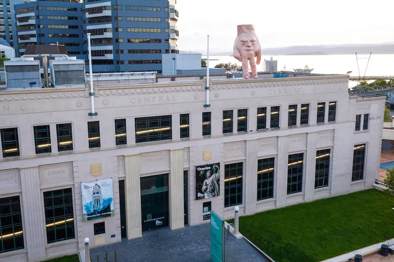 A large beige building with banners on the facade, labeled "Central Library." On the roof is a surreal sculpture of a large hand with a face. Modern glass buildings and water are visible in the background.