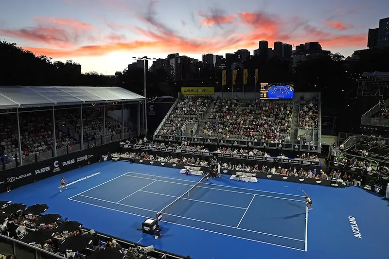 A wide-angle view of a blue tennis court during a match at sunset, with a large crowd in the stands and city buildings silhouetted against a colorful sky in the background.
