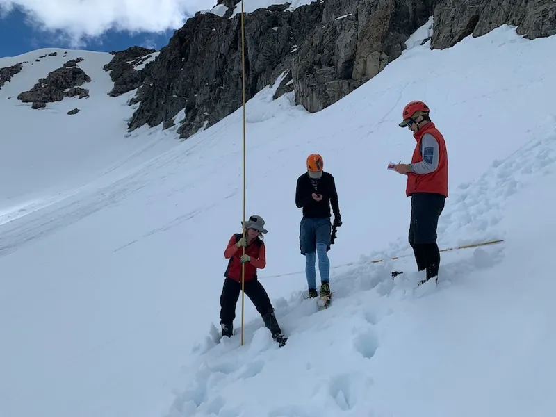 a group of people on a snowy mountain wearning helmets. one has stripey polypro and another is holding a long pole.