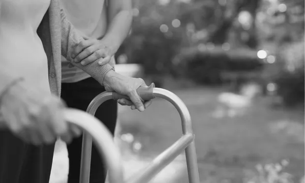 A close-up of an elderly person using a walker outdoors, with another person gently supporting them by holding their arm. The scene is in black and white.