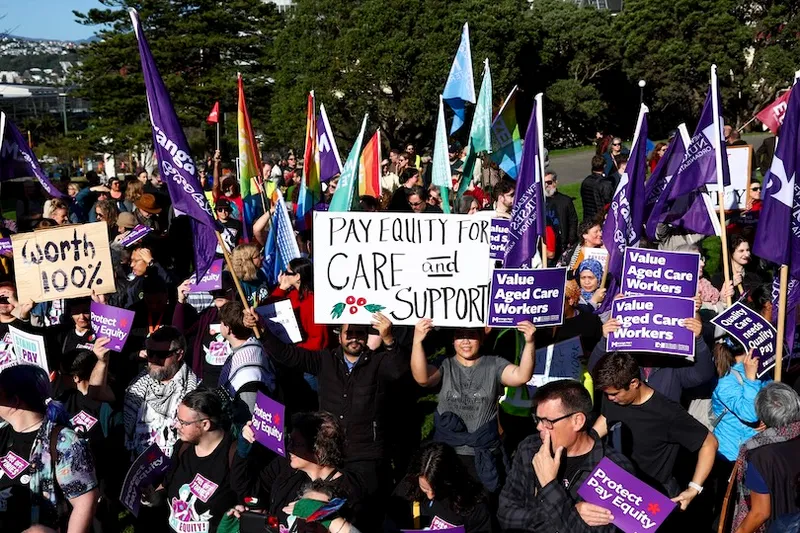 A large crowd of protesters holding signs and banners advocating for pay equity and support for aged care workers, gathered outdoors on a sunny day.
