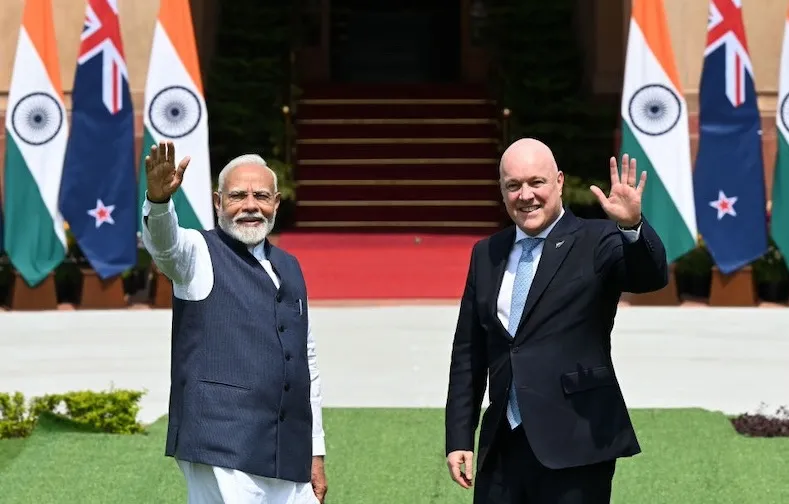 a white man and a brown man in a vest wave at the camera with the flags of New Zealand and India behind them