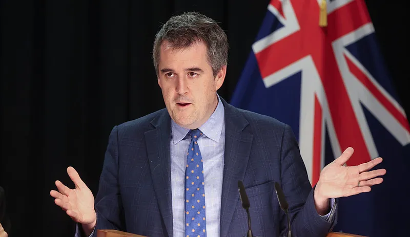 A man in a blue suit and polka dot tie speaks at a podium with his hands raised, standing in front of a partially visible flag featuring the Union Jack.