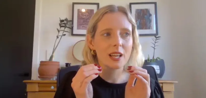 A woman with blonde hair and red nail polish gestures with her hands while speaking. She is seated indoors in front of a light-colored wall, with framed artwork and potted plants visible behind her.