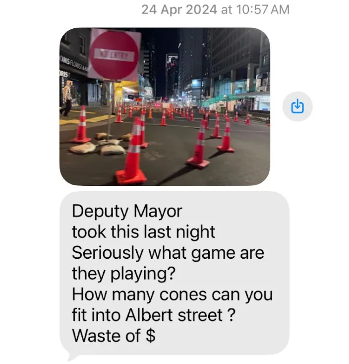 A city street at night is blocked by numerous orange traffic cones and a "no entry" sign. Below, a text message questions the excessive cones and calls it a waste of money.
