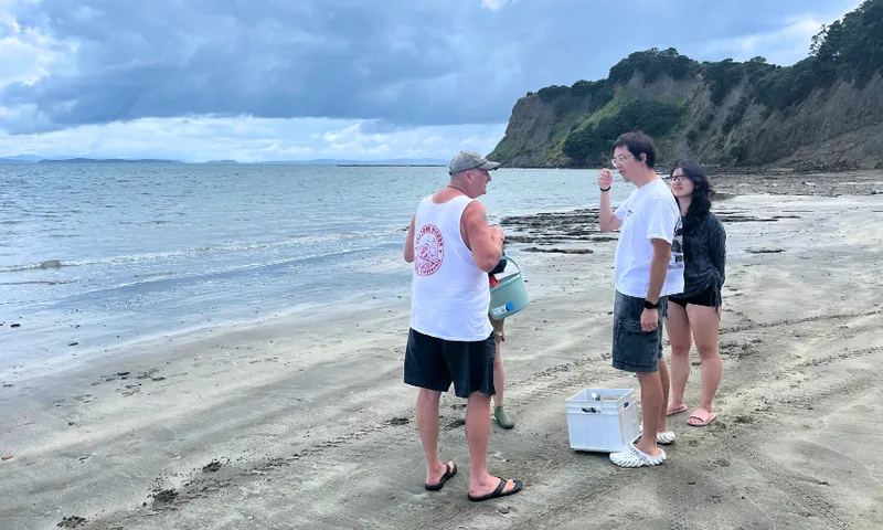 Four people stand and talk on a sandy beach near the water, with cliffs and trees in the background under a cloudy sky. One person holds a cooler, and another wears a white tank top and black shorts.