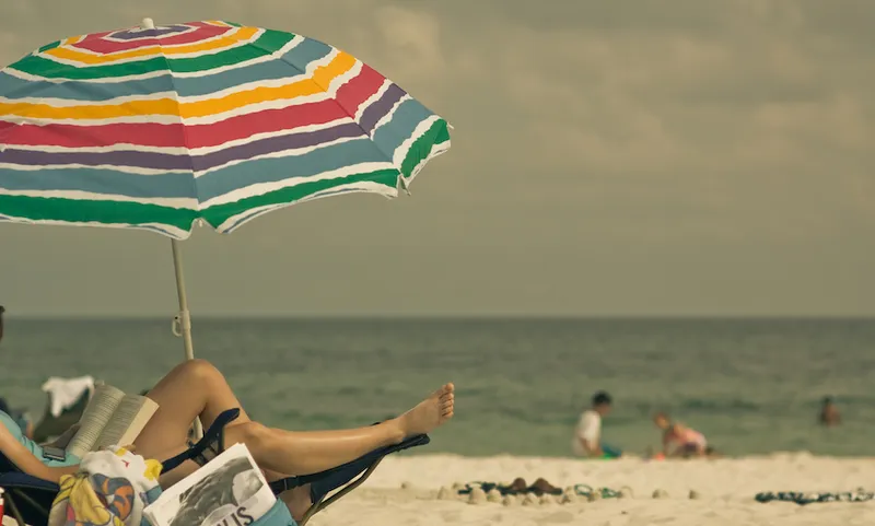 A beach, hot clouds, a rainbow sun umbrella, kids playing in the hazy distance at the water's edge. In the foreground we see part of a deckchair, and a person reading a book. We can't see their face.
