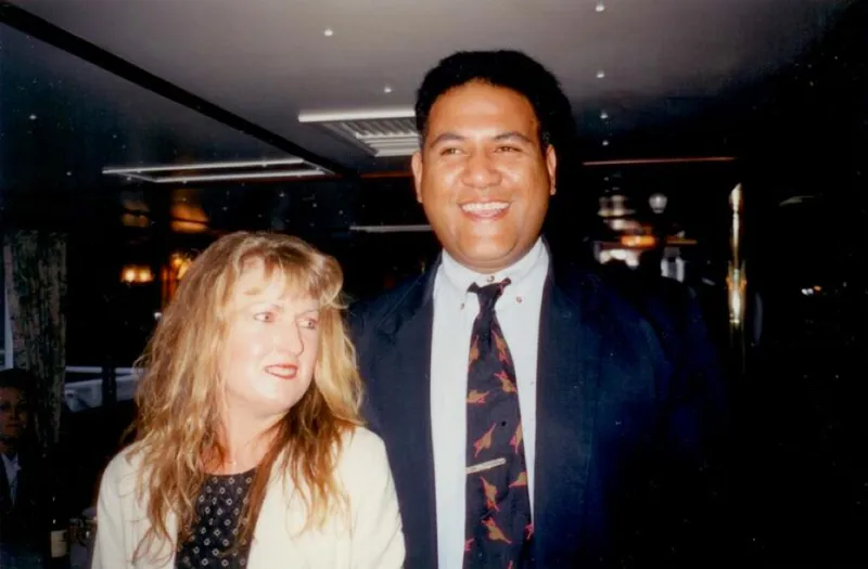 A woman with long blonde hair and a man in a suit and animal-patterned tie stand together indoors, smiling. The background shows a well-lit room with tables and people.