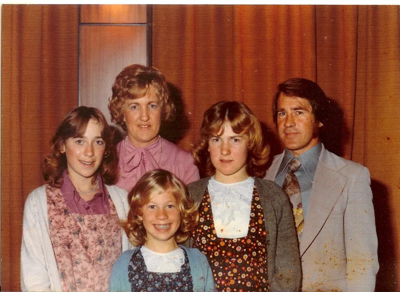 A vintage photo of a family of five posing indoors in front of brown curtains. Three girls stand in front, with an adult woman and man behind them. Everyone is dressed in 1970s-style clothing and smiling at the camera.