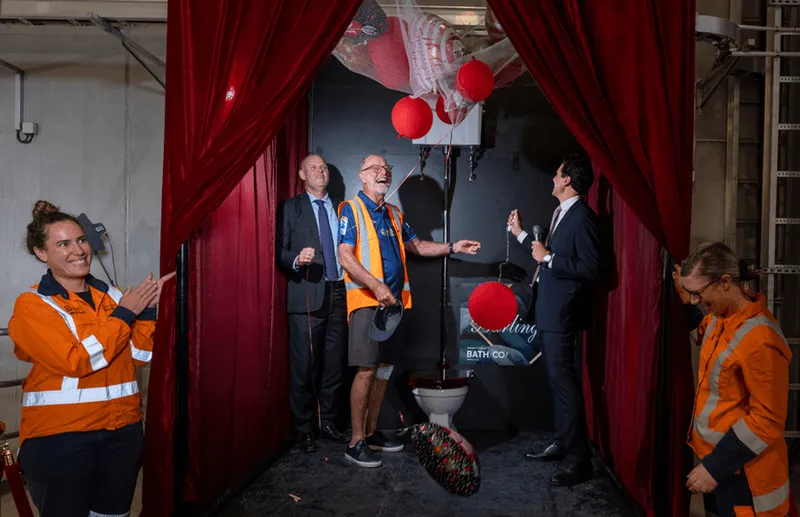 Several people in safety vests and suits laugh and celebrate in front of a stage with red curtains, balloons, and a toilet underneath a piñata-like object. One woman gestures toward the scene, while another looks down, smiling.