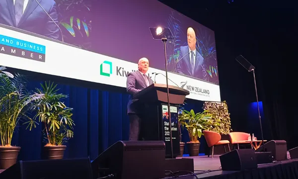 A man in a suit speaks at a podium on a stage with large screens displaying his image. The backdrop features plants, chairs, and business logos. The venue is set up for a formal event or conference.