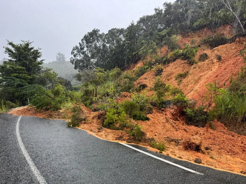 A landslide covers part of a wet road with red soil and debris during heavy rain, blocking one lane. Trees and greenery are visible on the roadside and hillside.