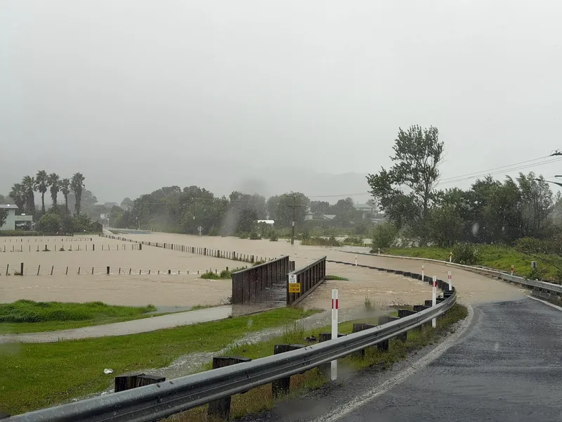 A flooded road and bridge are surrounded by rising water, with trees and a cloudy sky in the background. Rain is falling, and visibility is low, indicating severe weather and hazardous driving conditions.