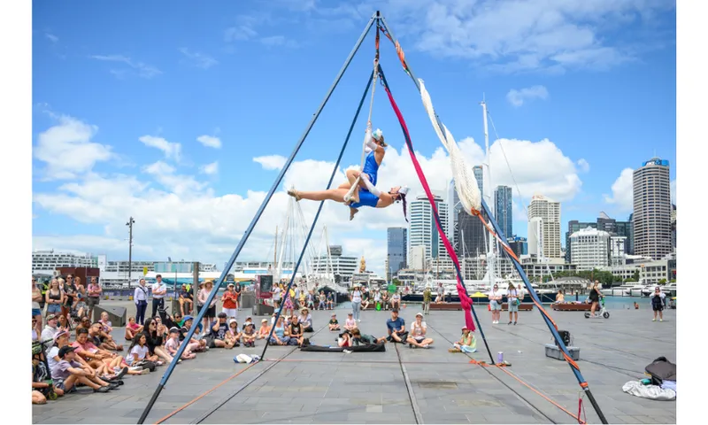 A photograph of circus performers set up outside on a waterfront space with a crowd watching.