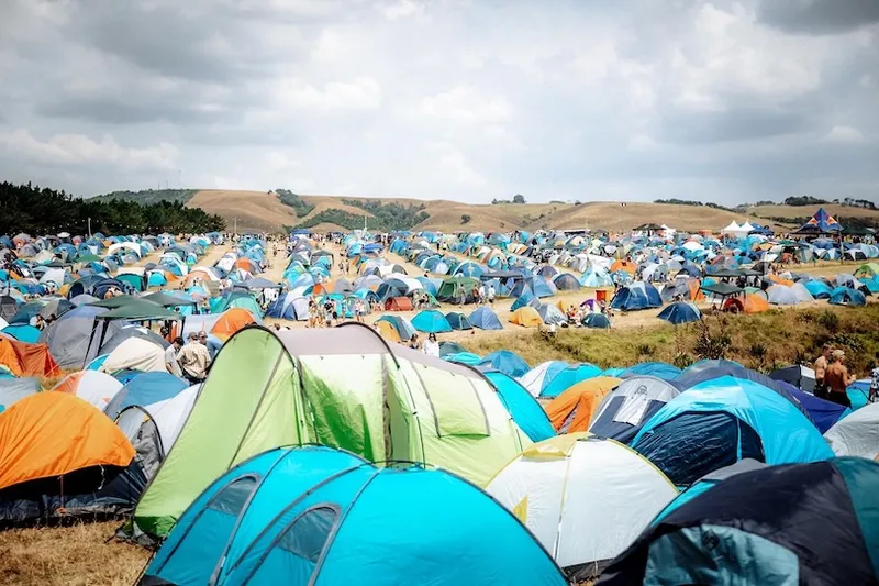 A large field filled with colorful tents under a cloudy sky, with rolling hills in the background and many people visible among the tents, suggesting a festival or large outdoor event.