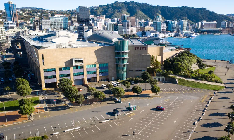 A large modern building with a cylindrical glass entrance sits beside a waterfront in a city with hills in the background; the adjacent parking lot is mostly empty under a clear sky.