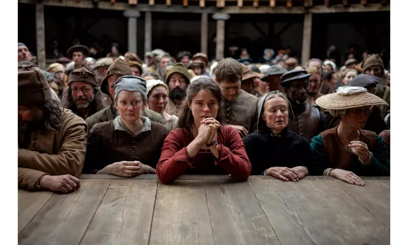 A photograph from the set of the movie Hamnet in which a crowd are standing in the pit of The Globe theatre and all look sombre.