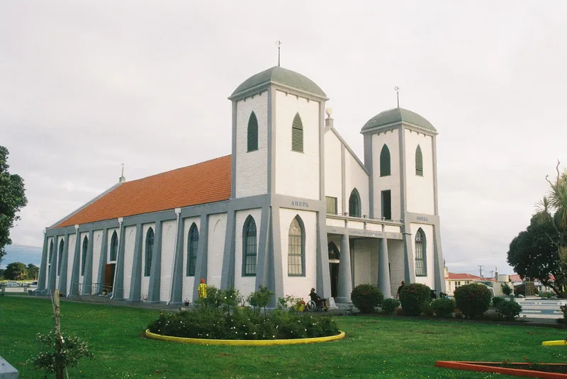 A white church with two tall towers, a red roof, arched windows, and green doors, sits on a well-kept lawn with a flowerbed in front. The sky is overcast and houses are visible in the background.