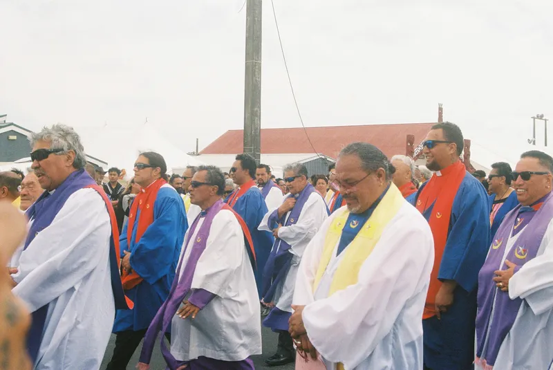 A group of people wearing colourful robes and sunglasses walk outdoors in a procession, some smiling. The robes are white, blue, red, and purple, and there are tents and a building in the background.
