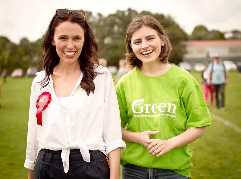 Two women smiling outdoors; one wears a white blouse with a red ribbon, and the other wears a green T-shirt with “Green Party of Aotearoa New Zealand” printed on it. People and trees are visible in the background.