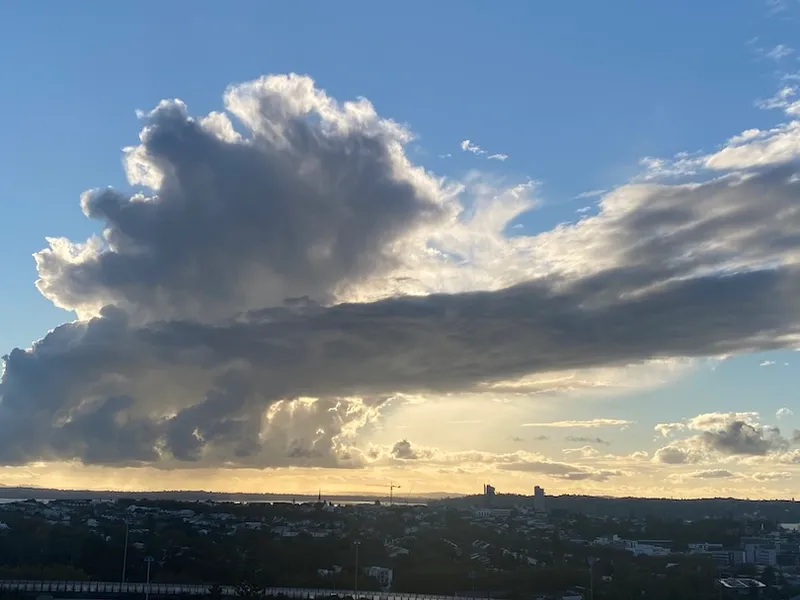 Large, dramatic clouds illuminated by the setting sun stretch across a blue sky above a silhouetted cityscape and distant water, creating a striking contrast between light and shadow.