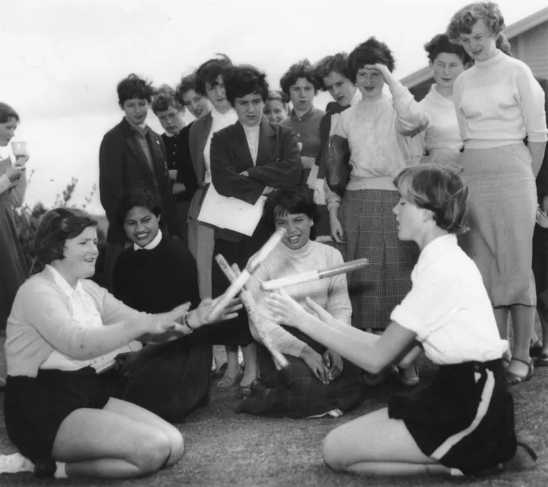 A group of young women watch as two girls, seated on the ground, play a game using sticks. The spectators are smiling and appear engaged with the activity, which takes place outdoors.