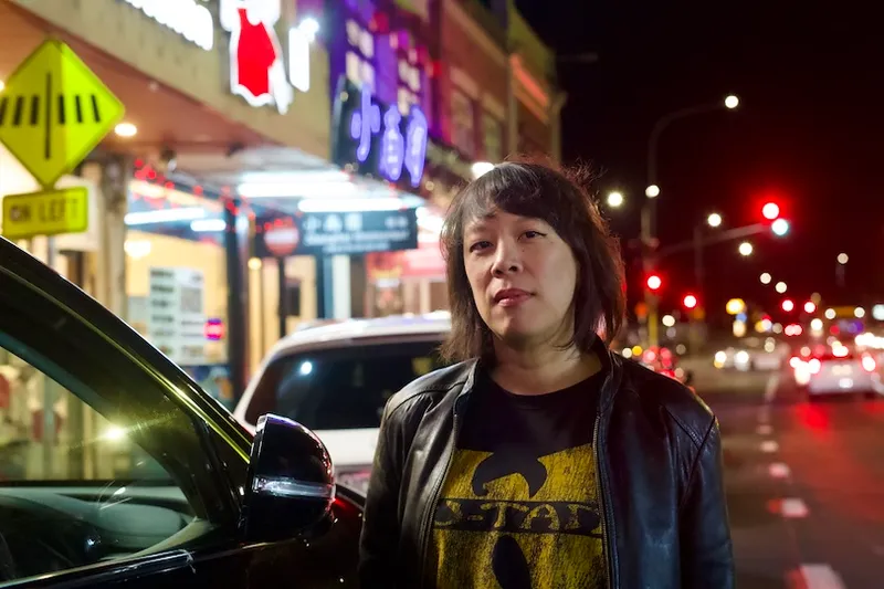 A person with medium-length dark hair wears a black leather jacket and a Wu-Tang Clan t-shirt, standing next to a car on a brightly lit city street at night. Neon signs and traffic lights are visible in the background.