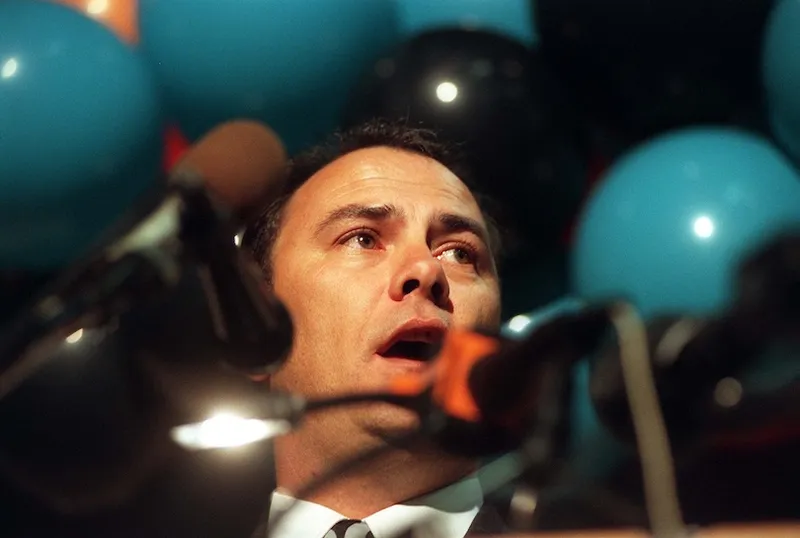 A man speaking into multiple microphones with blue and black balloons in the background.