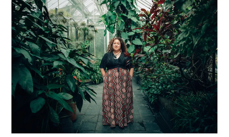 A photograph of a young Sāmoan woman standing in a greenhouse.