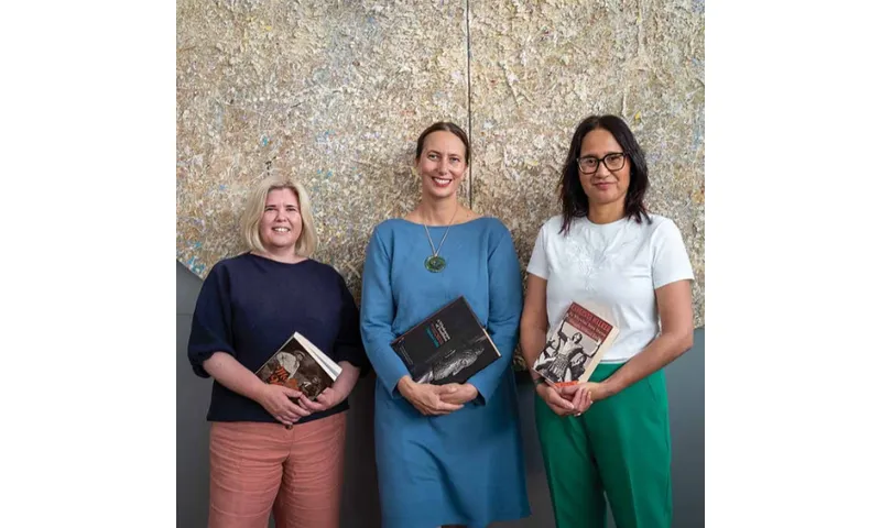 Three women standing side by side, each holding books in their hands and smiling.