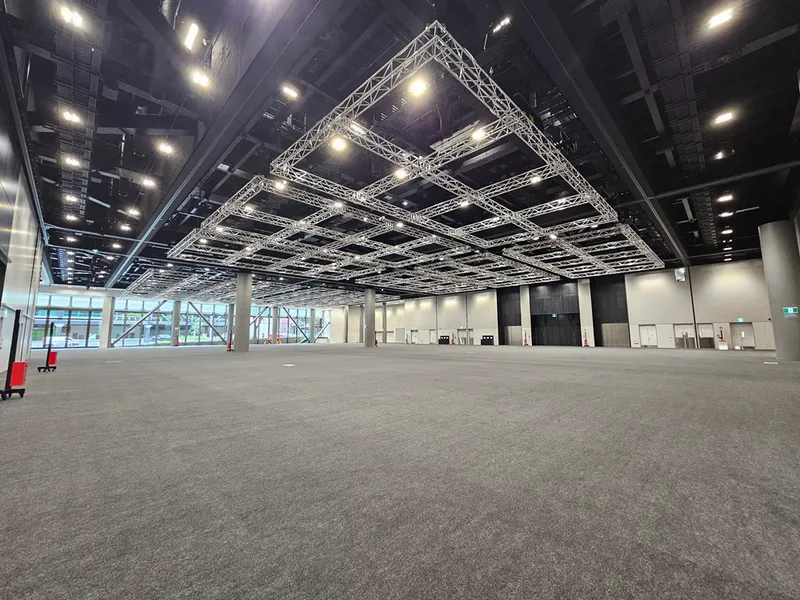 A large, empty convention hall with grey carpet, high ceilings, metal framework for lighting, and glass windows on one side letting in natural light. The space appears ready for an event setup.