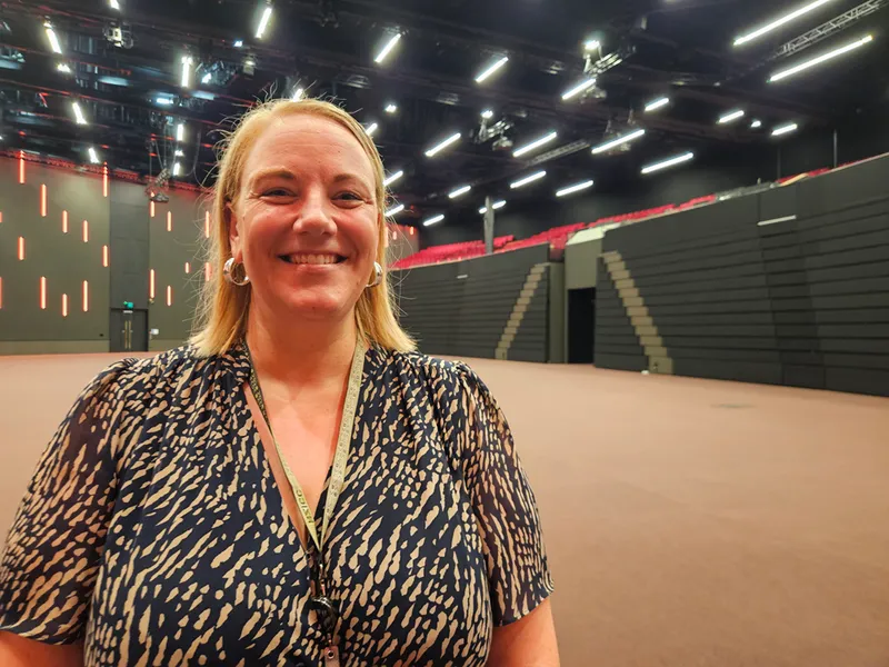 A woman with blonde hair, wearing a patterned blouse and lanyard, smiles while standing in a large, empty, modern auditorium with black walls and red seats in the background.