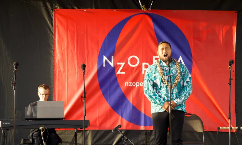 A Pasifika male singing into a microphone with a man on the left playing the keyboard in accompaniment. Behind them is a large banner for NZ Opera.