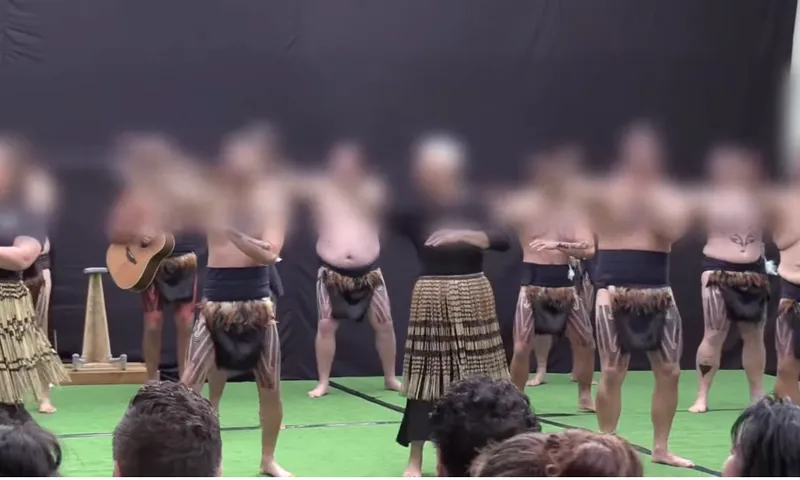 A group of people in traditional Māori attire perform a cultural dance on a stage with audience members visible in the foreground. The background is plain and dark.