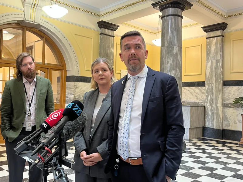 a tall man in a blue tie and a shorter blonde woman wearing a grey blazer look sideways with a bevy of microphones in front of them and distinctive black and white parliamment tiles behind them.