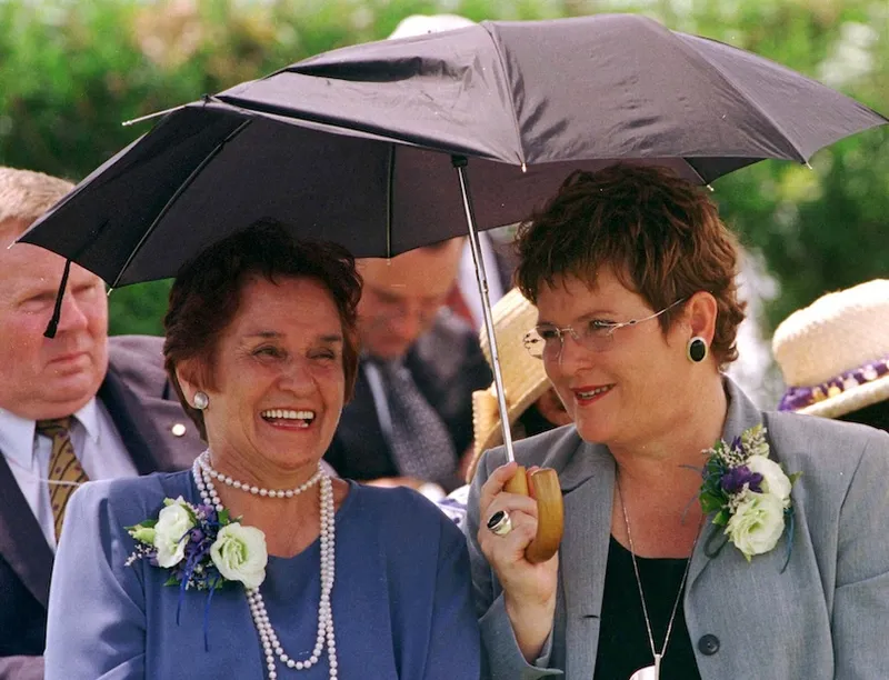 Two women dressed formally sit together, smiling and talking under a black umbrella at an outdoor event. Both wear flower corsages and pearl necklaces. Other people are visible in the background.