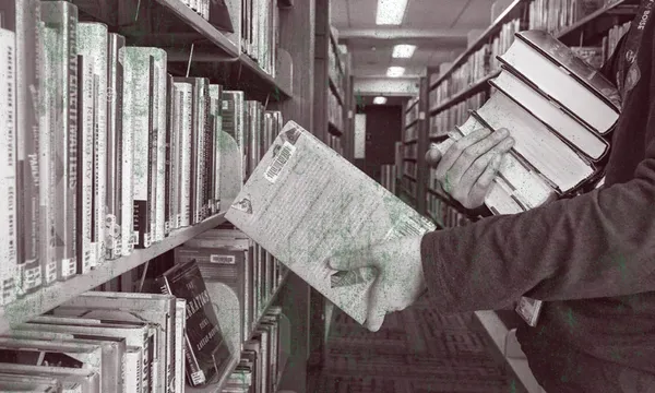 A person stands between library shelves, holding a stack of books in one arm while reaching with the other hand to pull a book from the shelf. The scene is in black and white.
