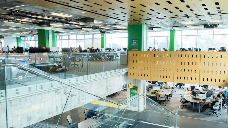 A modern, open-plan office space with two levels, featuring glass walls, green columns, and people working at desks and computers. A staircase connects the floors, and large windows let in natural light.