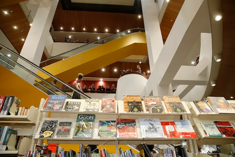 Bookshelves filled with graphic novels and magazines in the foreground of a modern, multi-level library with people visible on upper floors and warm lighting throughout the space.