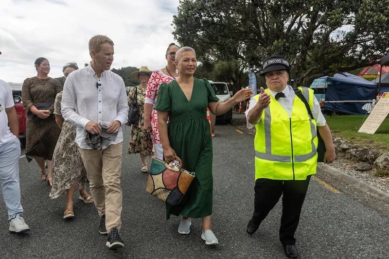 A group of people walks outdoors. A woman in a green dress talks with a man in a light shirt, while a person in a bright yellow safety vest and hat leads them along a paved path. Other people and tents are in the background.