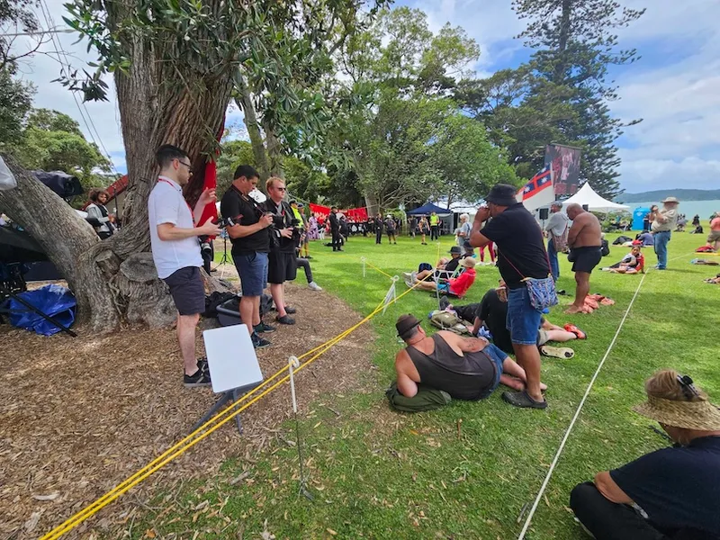 A group of people gather in a park with trees and tents for an outdoor event. Some stand by a large tree holding papers, while others sit or lie on the grass, and a yellow rope divides the area.