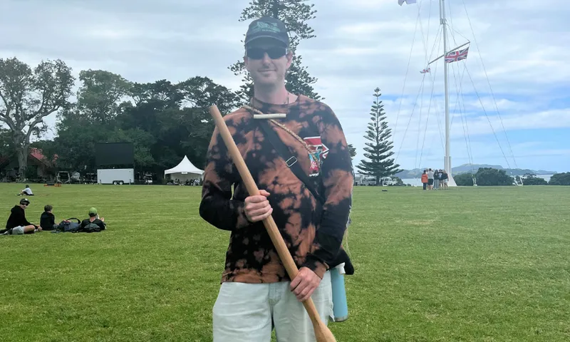 A man wearing sunglasses, a cap, and a tie-dye shirt stands on a grassy field holding a long wooden stick. In the background, groups of people sit on the grass near a tall flagpole and trees.