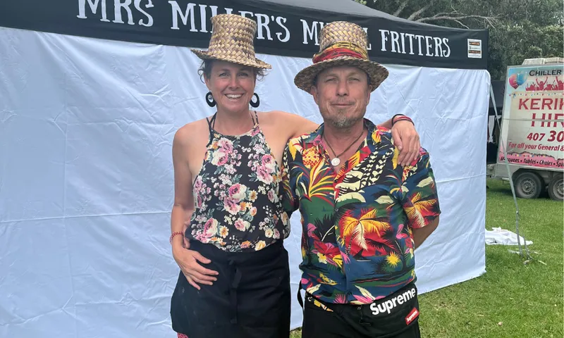 Two people wearing straw hats and aprons stand smiling in front of a banner that reads "MRS MILLER'S MUSSEL FRITTERS." They are outdoors, possibly at a food stall or market event.